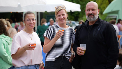 Drei Besucherinnen und Besucher blicken mit kühlen Getränken in die Kamera beim Albertinen-Sommerfest für die Mitarbeitenden aus den unterschiedlichen Einrichtungen der Immanuel Albertinen Diakonie am Standort Schnelsen auf dem Platz vor der Kirche am Albertinen Krankenhaus.