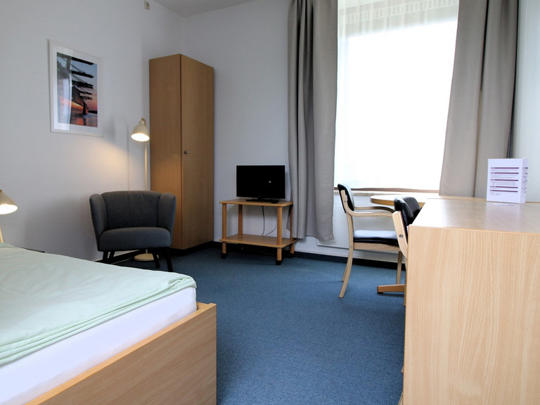 Blick in ein helles Gästezimmer mit blauem Teppich, Bett, Tisch, Sessel und einem großen Fenster der Albertinen Akademie in Hamburg für die Unterkunft bei Tagungen, Fortbildungen, Weiterbildungen im Gesundheitswesen sowie zu touristischen Buchung.