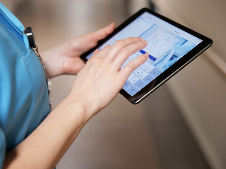 Nahaufname eines Tablets in Verwendung durch eine Pflegekraft in blauer Funktionskleidung auf einem Stationsflur für Webinare und Online-Fortbildungen der Albertinen Akademie Hamburg