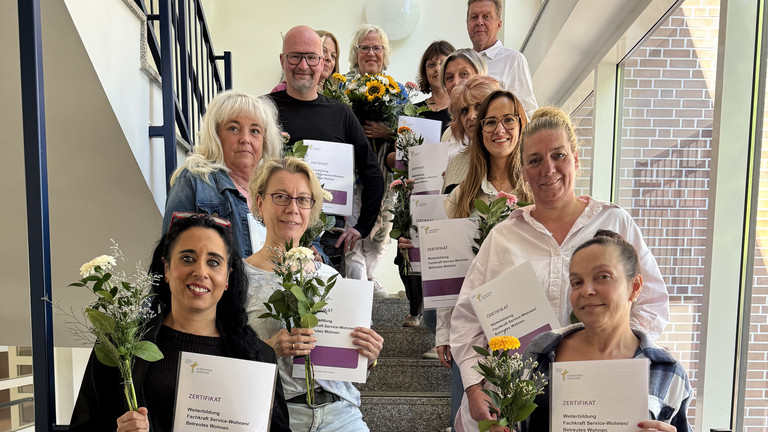 Gruppenfoto des 11. Jahrgangs der beruflichen Weiterbildung Fachkraft Service-Wohnen / Betreutes Wohnen mit Abschlusszertifikaten und Blumensträußen auf dem Treppenaufgang
