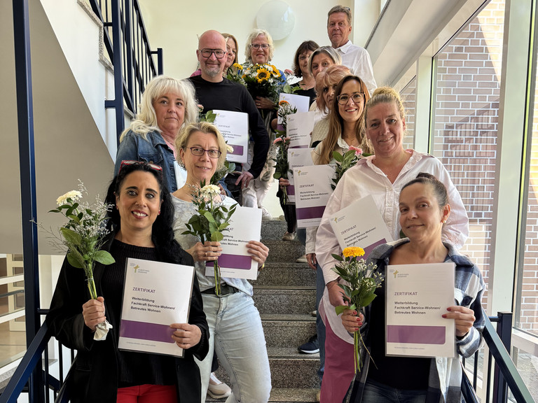 Gruppenfoto des 11. Jahrgangs der beruflichen Weiterbildung Fachkraft Service-Wohnen / Betreutes Wohnen mit Abschlusszertifikaten und Blumensträußen auf dem Treppenaufgang