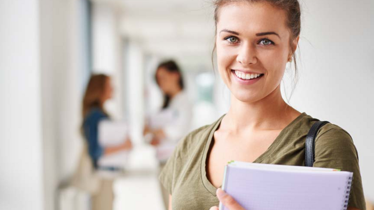 Eine Teilnehmerin blickt auf einem Gang mit weiteren Menschen mit einem Laptop in der Hand lächelnd in die Kamera als Titelmotiv zum Kursprogramm 2027 mit Seminaren, Fort- und Weiterbildungen für das Gesundheitswesen der Albertinen Akademie in Hamburg-Schnelsen.
